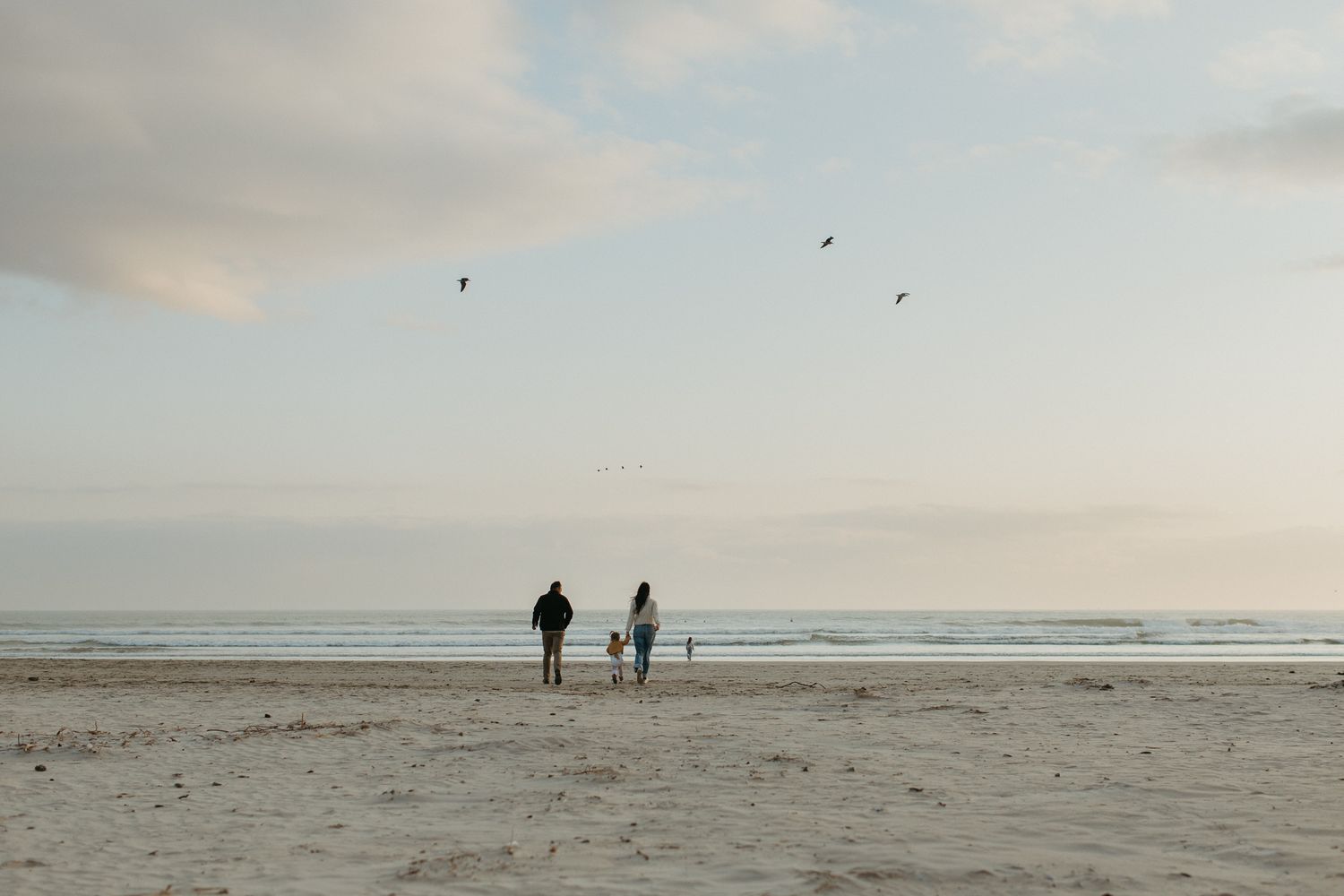 Two adults and a small child stand on a wide sandy beach as birds fly overhead beneath a soft pastel evening sky.