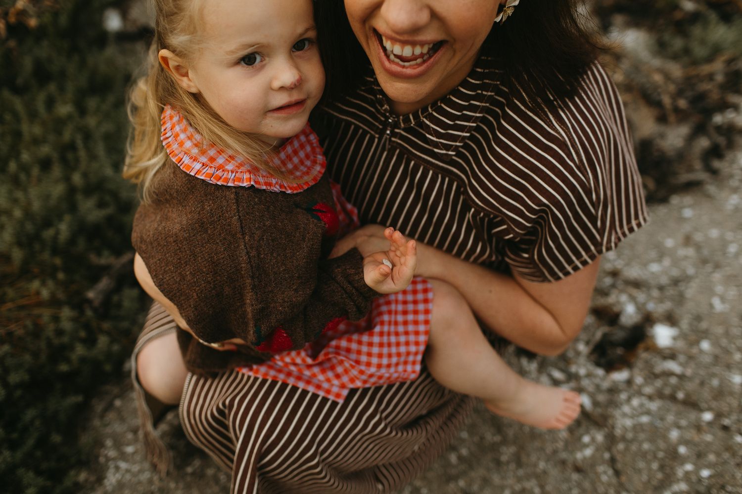 Smiling mother in a striped dress holding a toddler in a brown jacket and plaid dress outdoors.