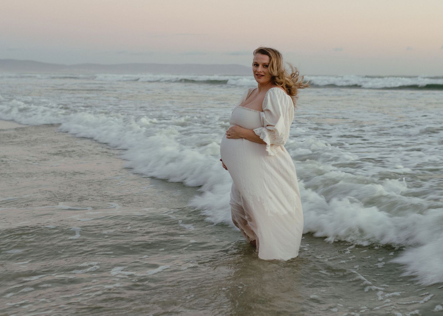 A pregnant woman in a flowing white dress stands in shallow ocean waves at dusk, holding her belly and smiling.