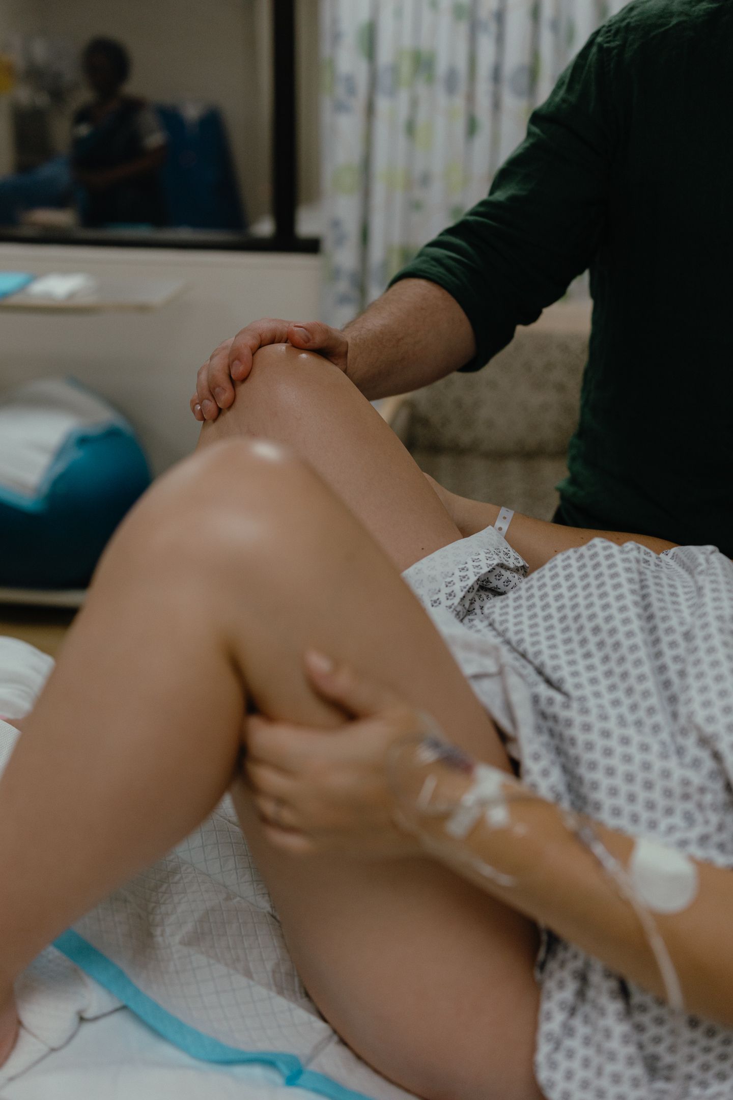 Person in a hospital gown with an IV in the hand lies in a hospital bed while a support person in green holds their knee.