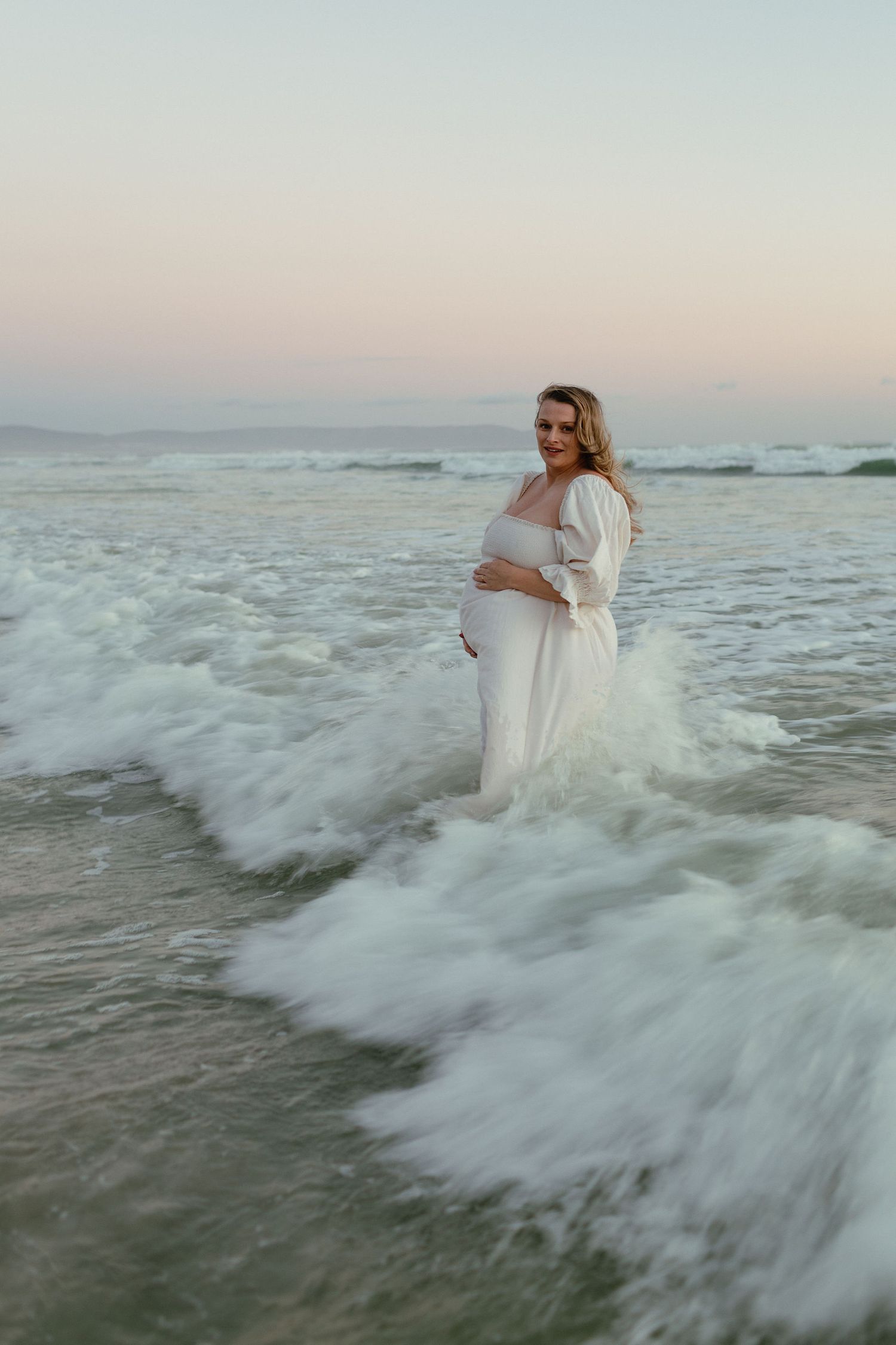A smiling pregnant woman in a white dress holds her belly while standing in ocean waves at a soft pastel sunset.