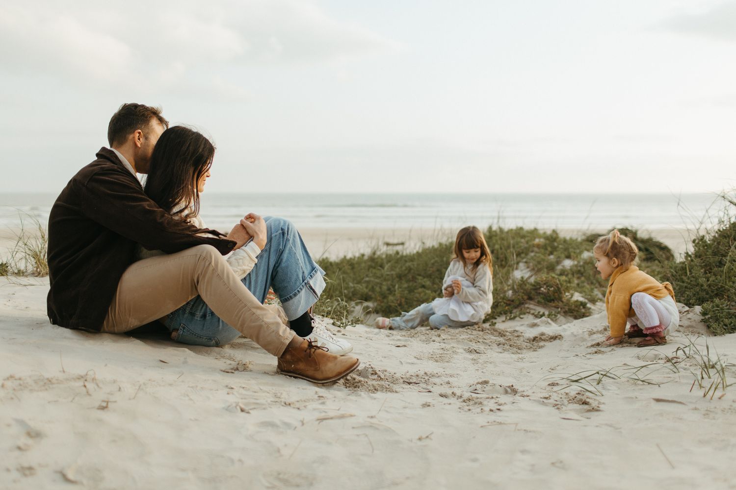 Couple sitting close together on a sandy beach while two young children play in the sand near the ocean at sunset.