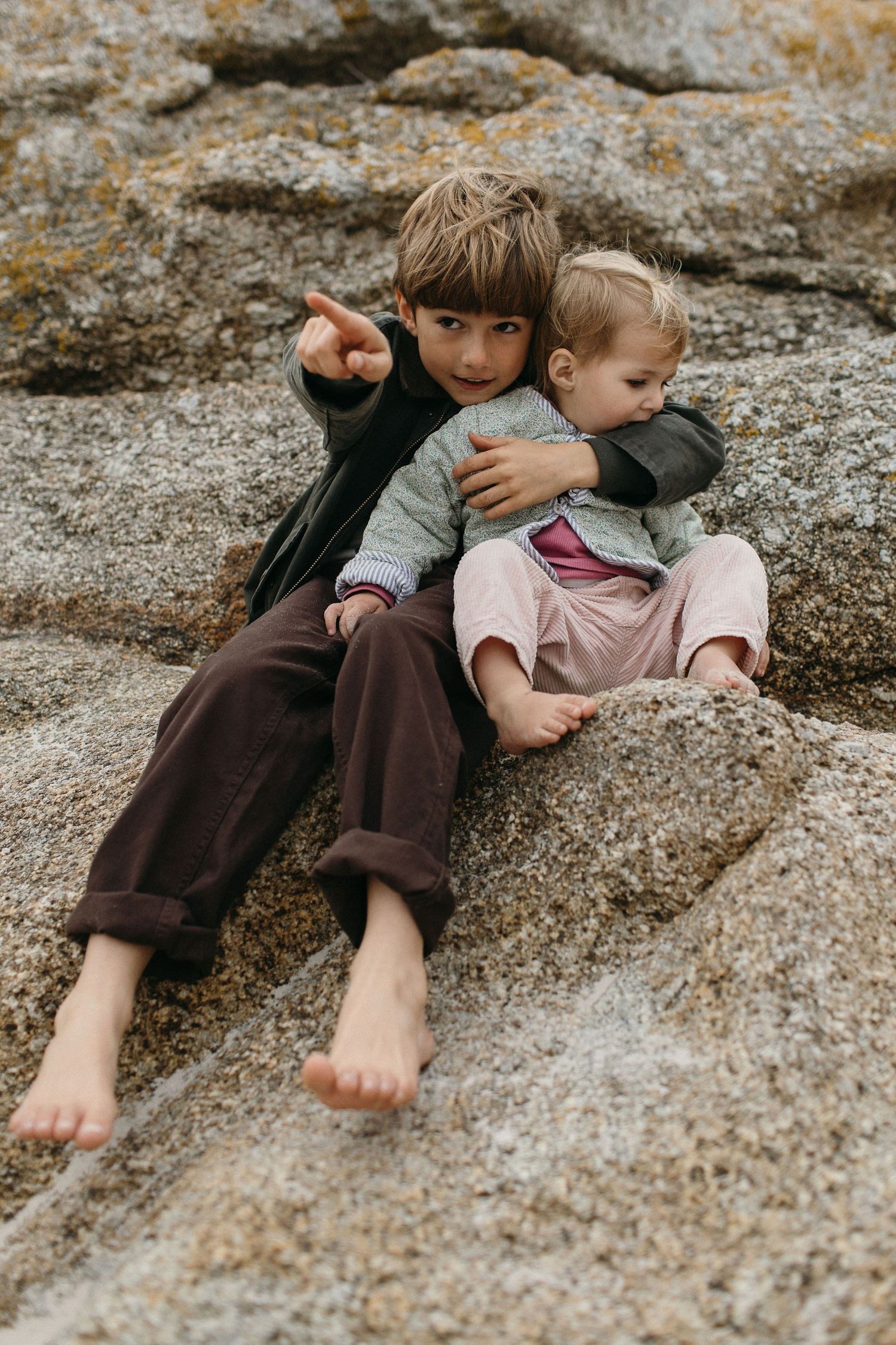 Two young brothers sit close together on a large granite boulder, one pointing into the distance while hugging his younger sibling.