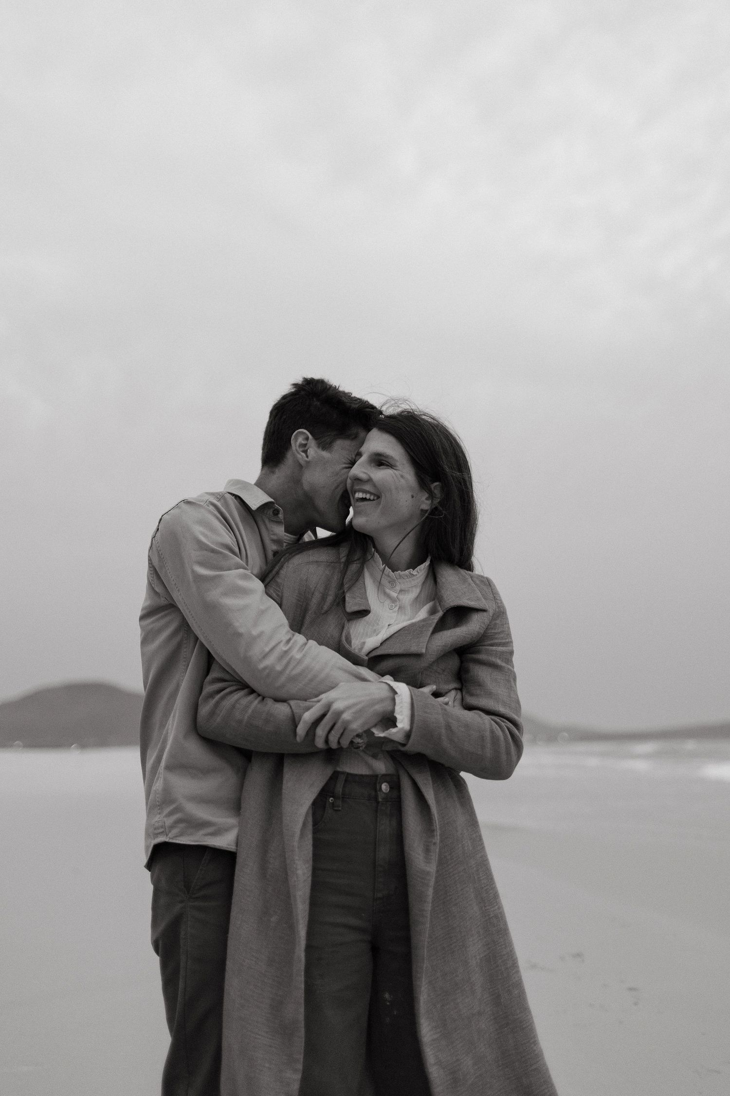 Black and white photo of a laughing couple embracing on a misty beach, the man kissing the woman's cheek.