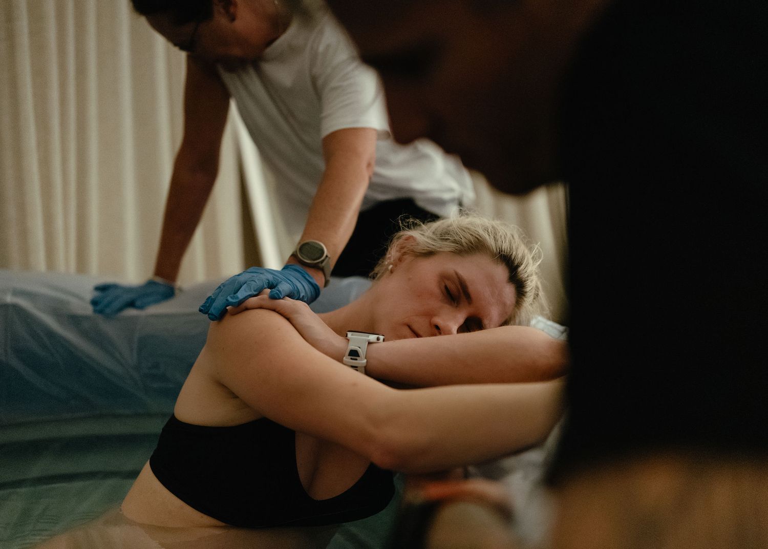 A woman in a black sports bra leans forward on a bed during labor as a birth partner rubs her back and another person watches nearby.