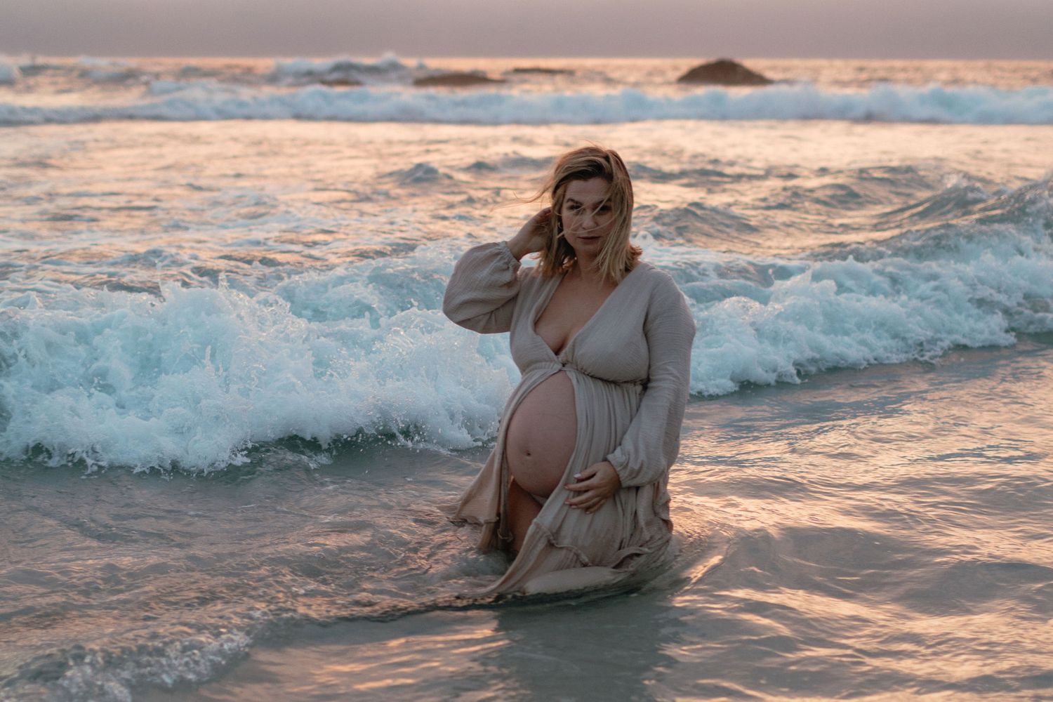 Pregnant woman in a flowing cream wrap sitting in shallow ocean waves at sunset during a maternity photo session.