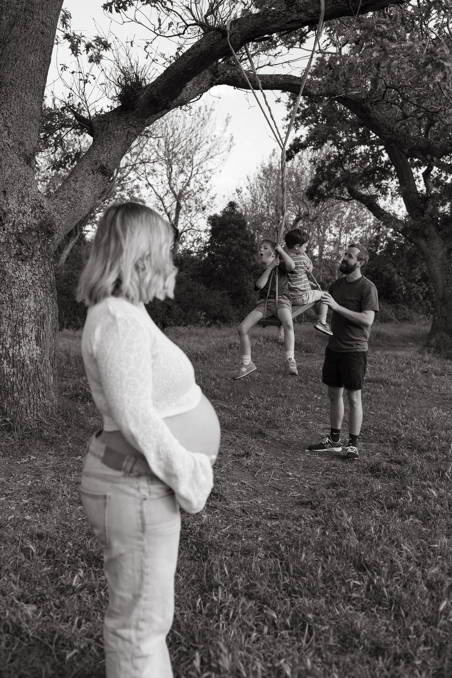 Black and white photo of a pregnant woman standing in a park while her husband pushes two children on a tree swing.