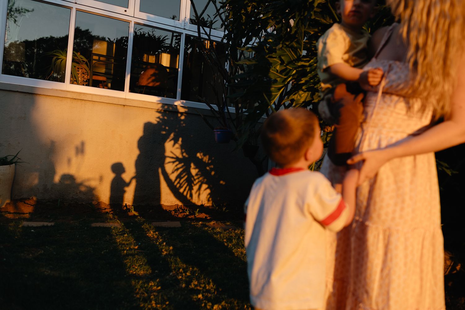 A little boy in a white shirt stands beside a woman in a cream dress as golden sunset light casts shadows on the garden wall.