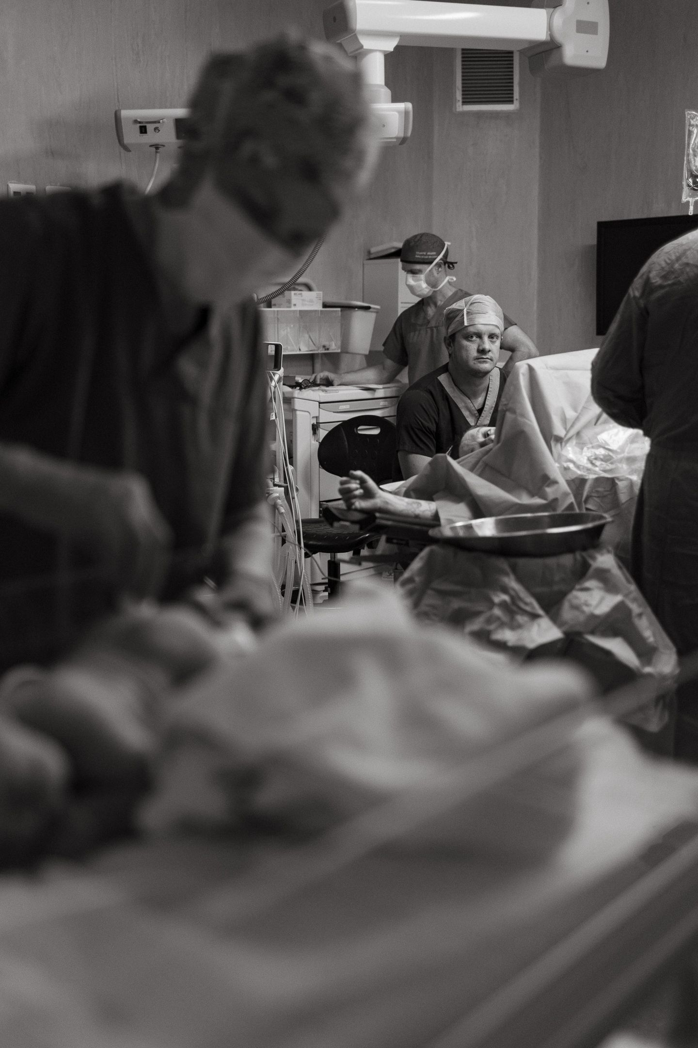 Black and white photo of surgical staff in scrubs and masks in an operating room during a cesarean birth, with a seated man watching.