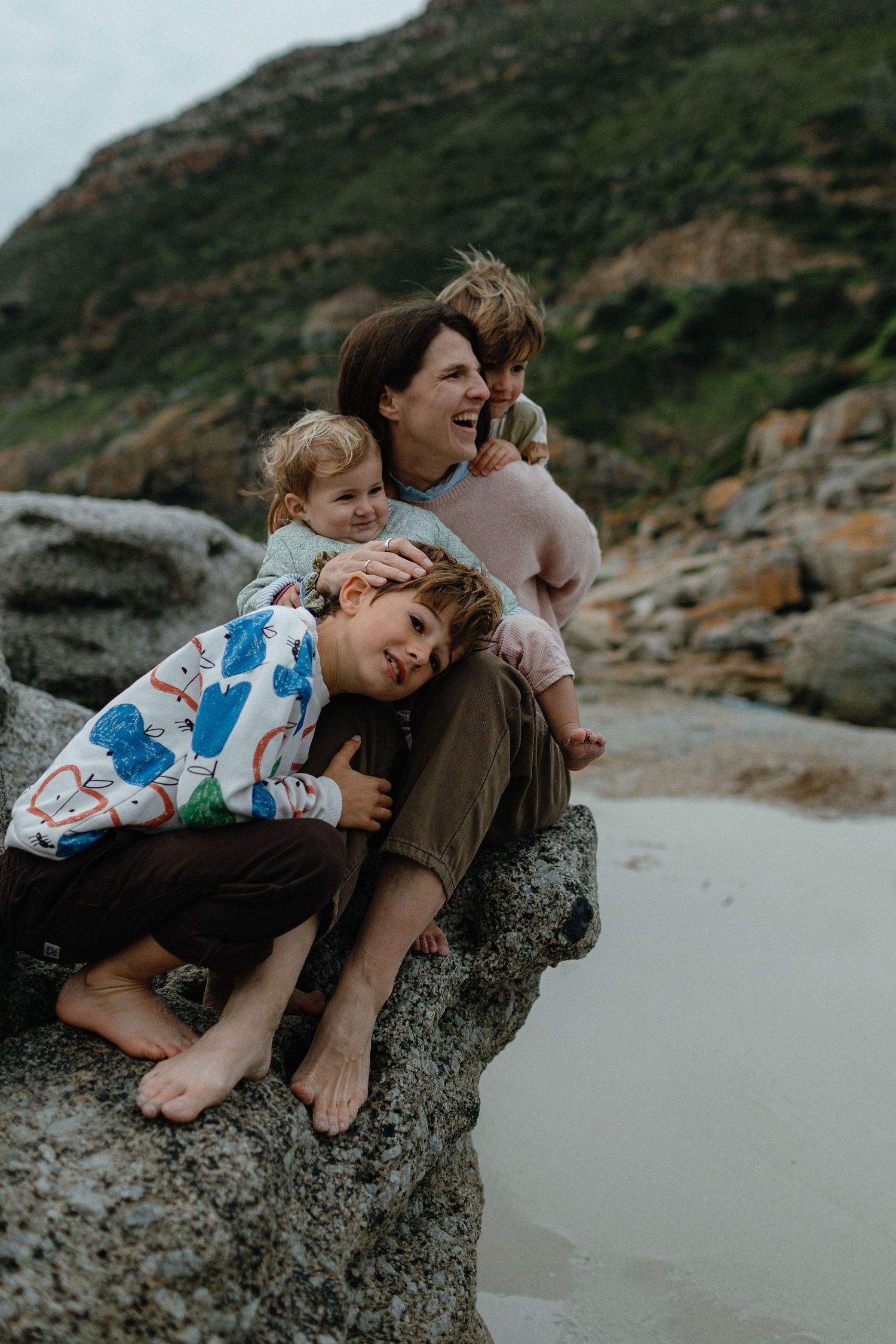 A smiling mother and three young children cuddle together on a coastal boulder with green mountains and a sandy beach behind them.