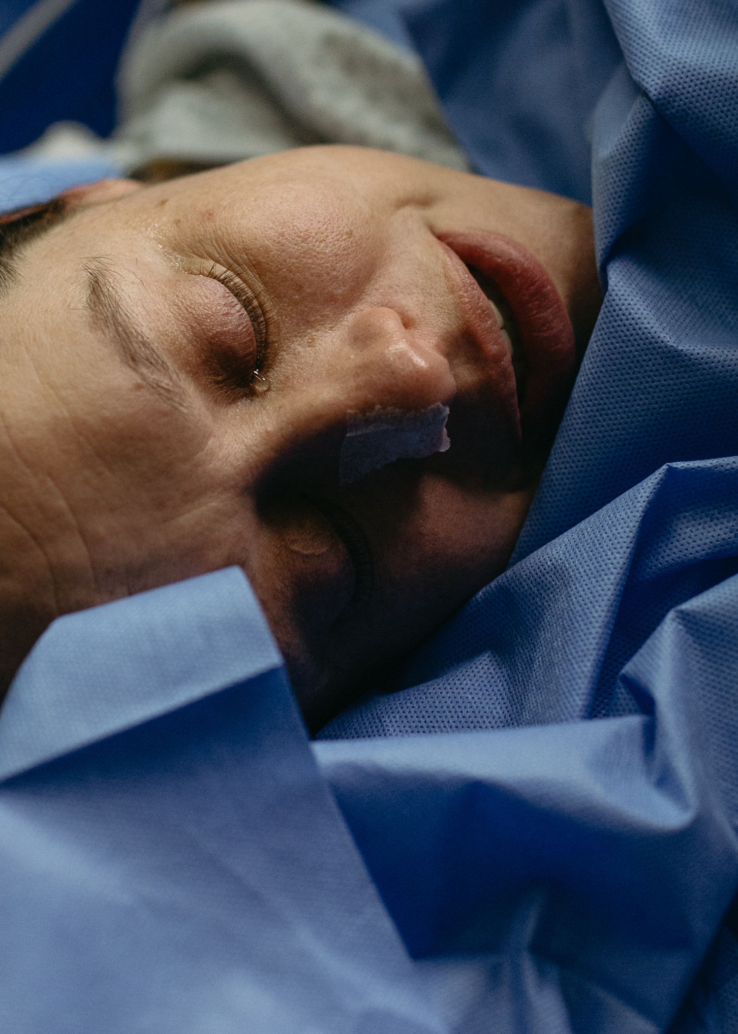 Close-up of a woman resting with closed eyes, wrapped in blue surgical drapes in a hospital delivery room.
