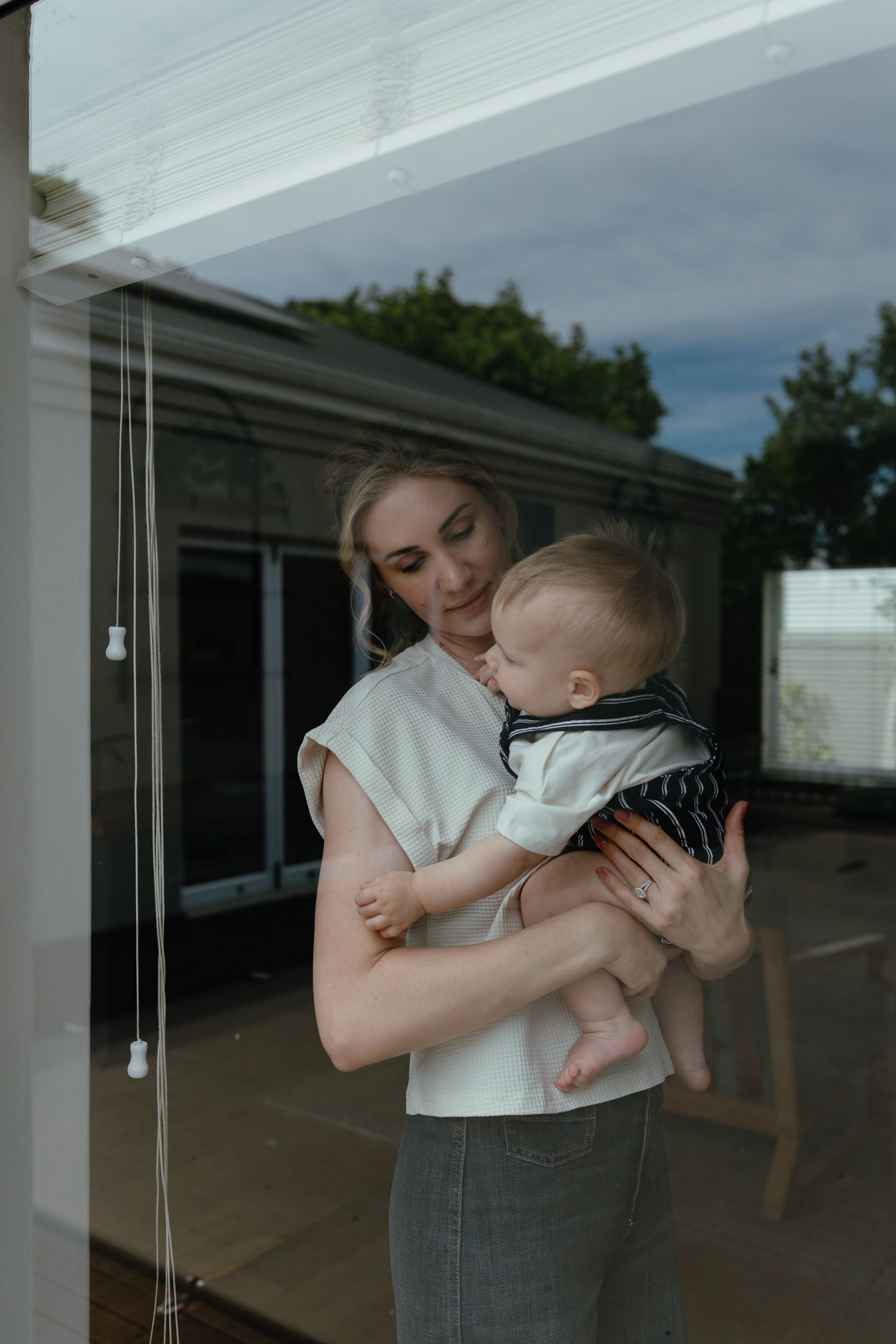A woman in a white sleeveless top holds a barefoot baby while standing behind a glass window.