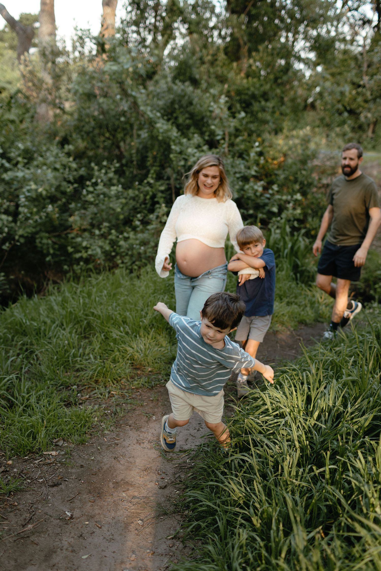 Pregnant mother walks a leafy trail with her husband and two young sons, smiling and playing in a joyful family outdoor moment.