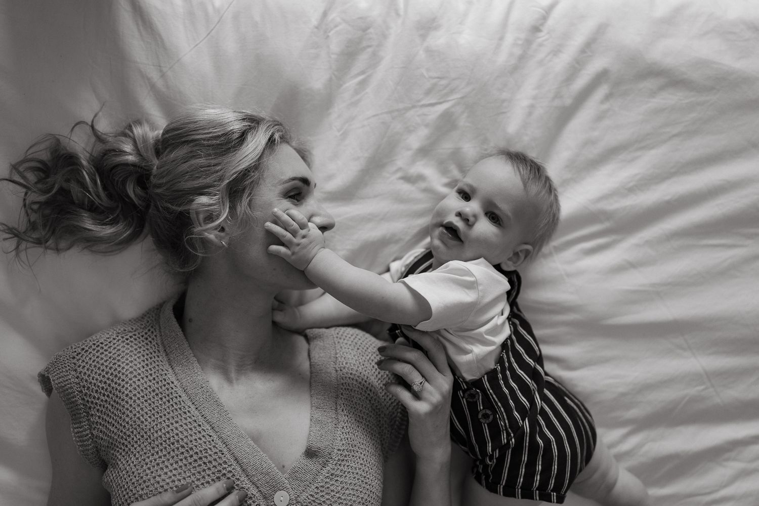 Black and white photo of a baby touching his mother's face as she lies on a bed with her hair spread across white sheets.