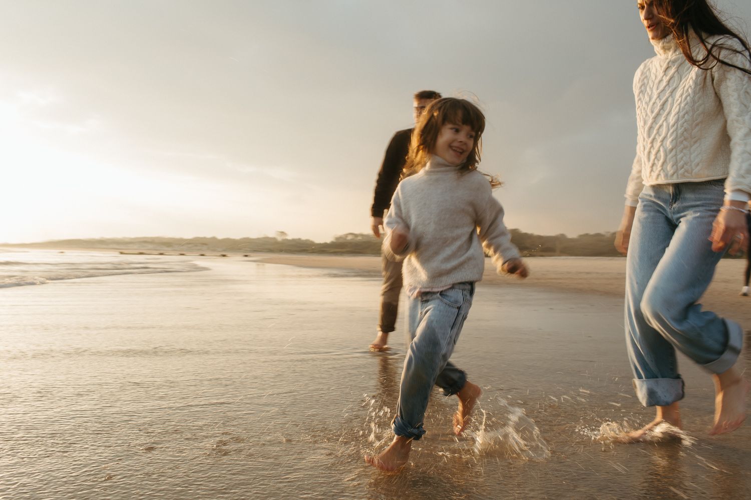 Smiling girl in a gray sweater and jeans runs through shallow ocean water at golden hour as her family follows behind.