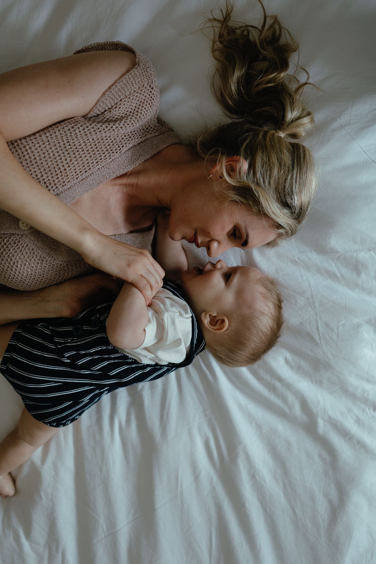Overhead view of a mother and baby lying nose to nose on white bed linens in a tender, quiet moment.