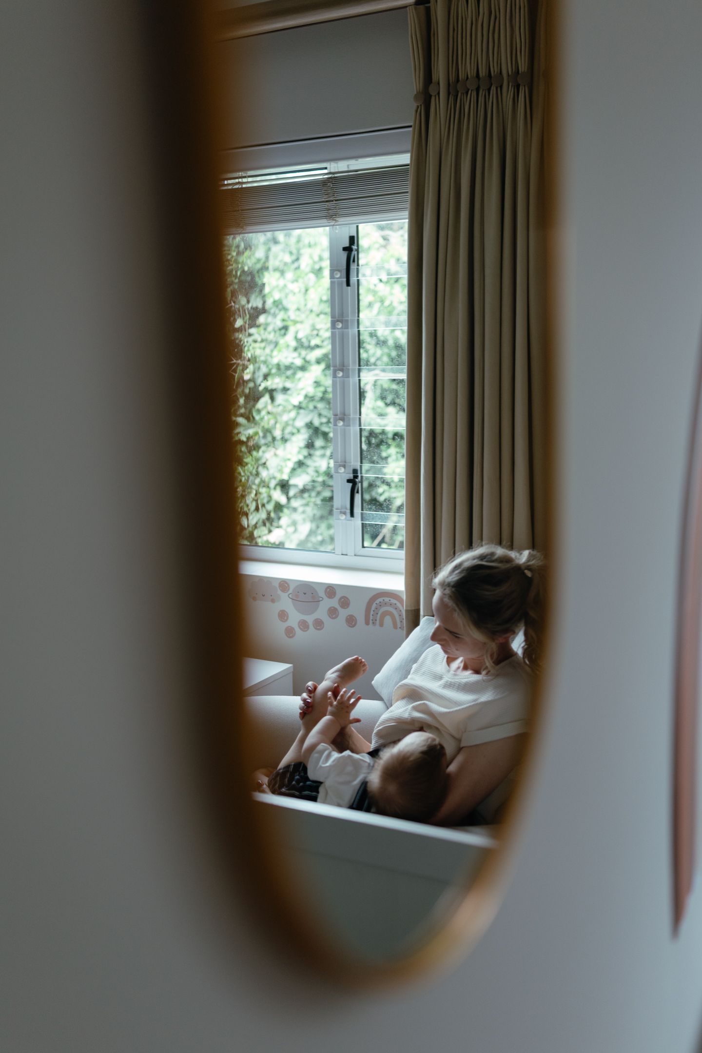 Mirror reflection of a mother cuddling her baby on a nursery chair beside a bright window with curtains.