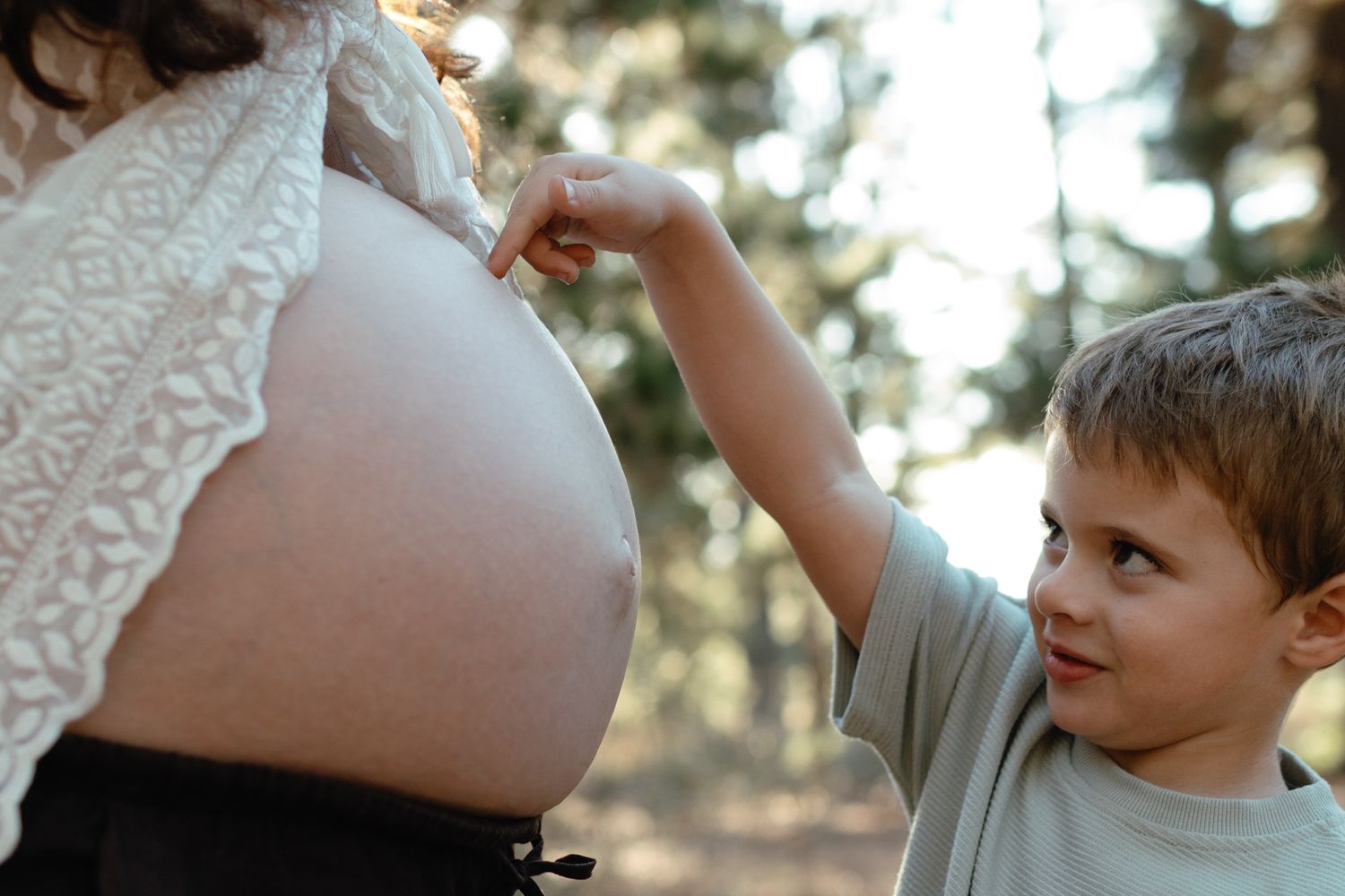 Curious young boy gently touches a pregnant woman's bare belly in a sunlit outdoor setting.