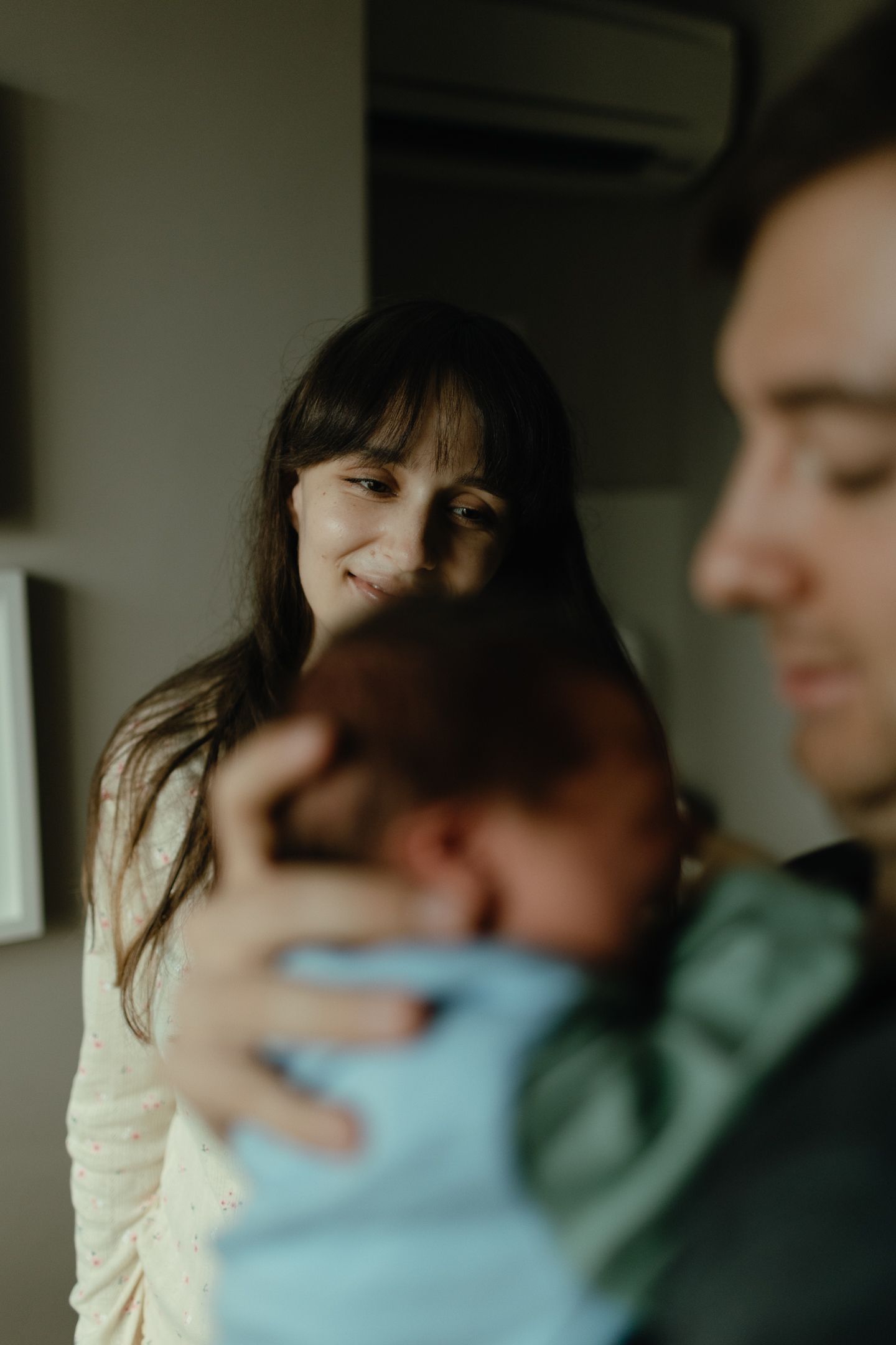 Smiling mother gazes lovingly at her newborn baby cradled in a blurred father's arms in a softly lit room.
