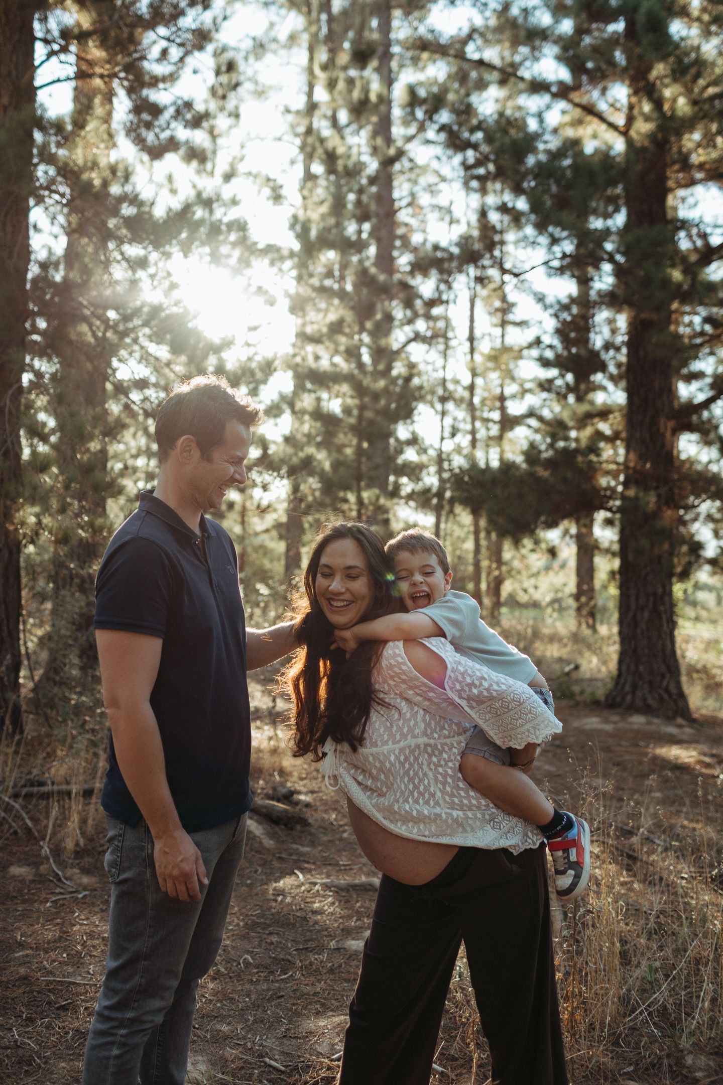 Happy family of three with a pregnant mother laughing and carrying her son beside her husband in a sunlit pine forest.