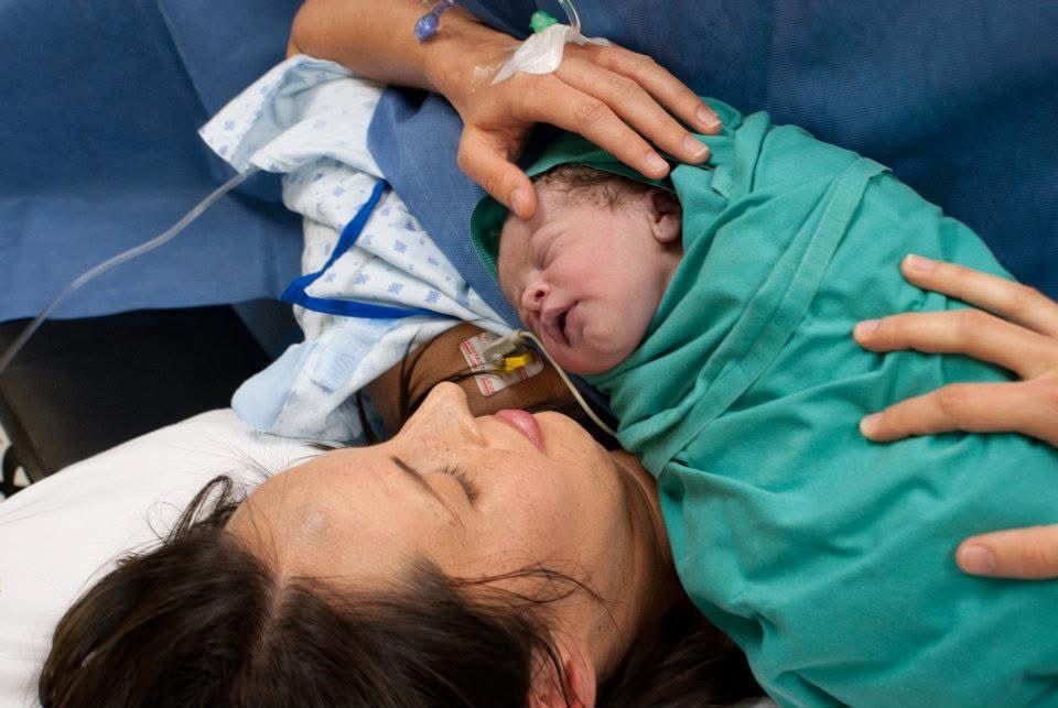 A new mother lies back and rests her cheek against her newborn baby, who is wrapped in a green blanket and held by a hand.