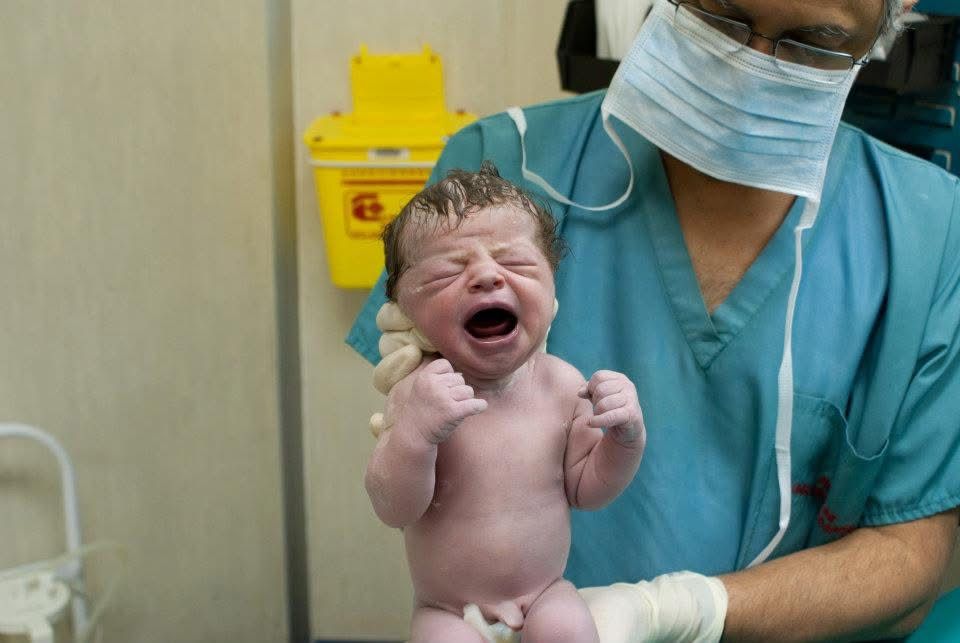 A masked doctor in blue scrubs holds up a crying, wide-eyed newborn baby in a hospital delivery room.