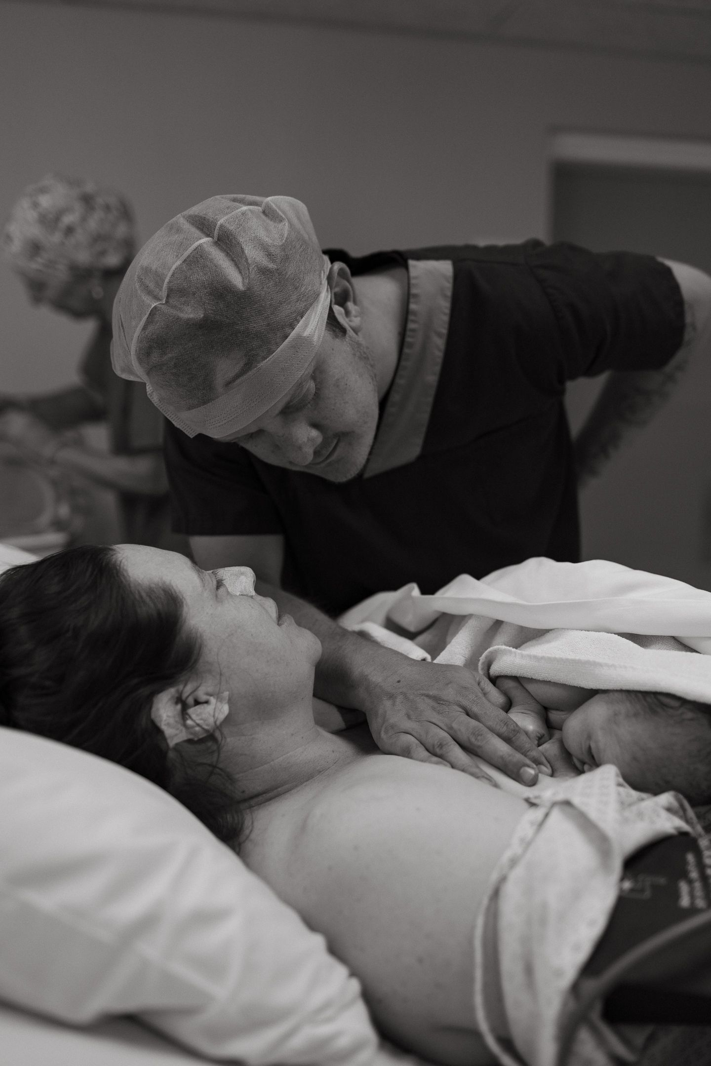 Black and white image of a smiling mother in a hospital bed holding her newborn while a nurse in a scrub cap leans in close.