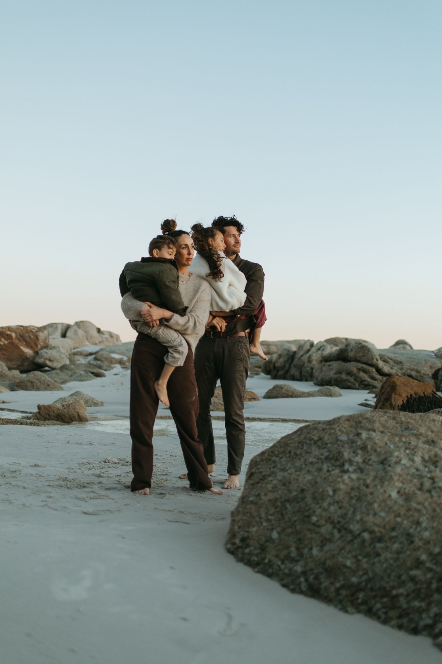 Parents hold their two children close while standing on a sandy beach beside large rocks at dusk.