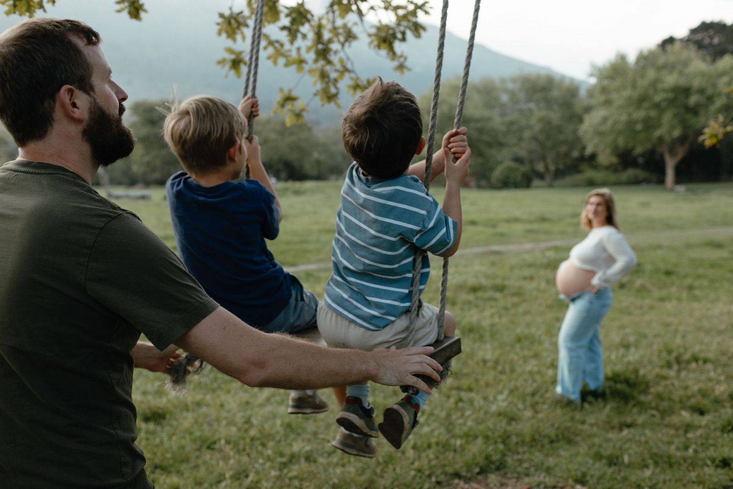 Father pushes two boys on a rope swing in a grassy field while a pregnant mother watches from the background.