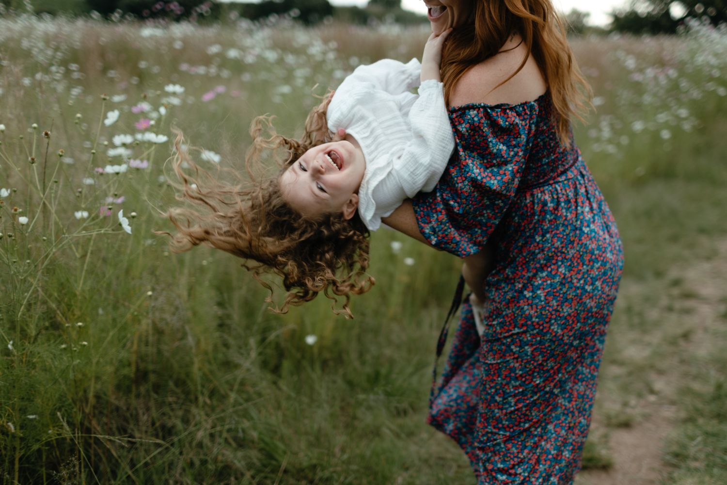 Laughing mother swings her daughter through the air in a field of tall grass and white wildflowers.