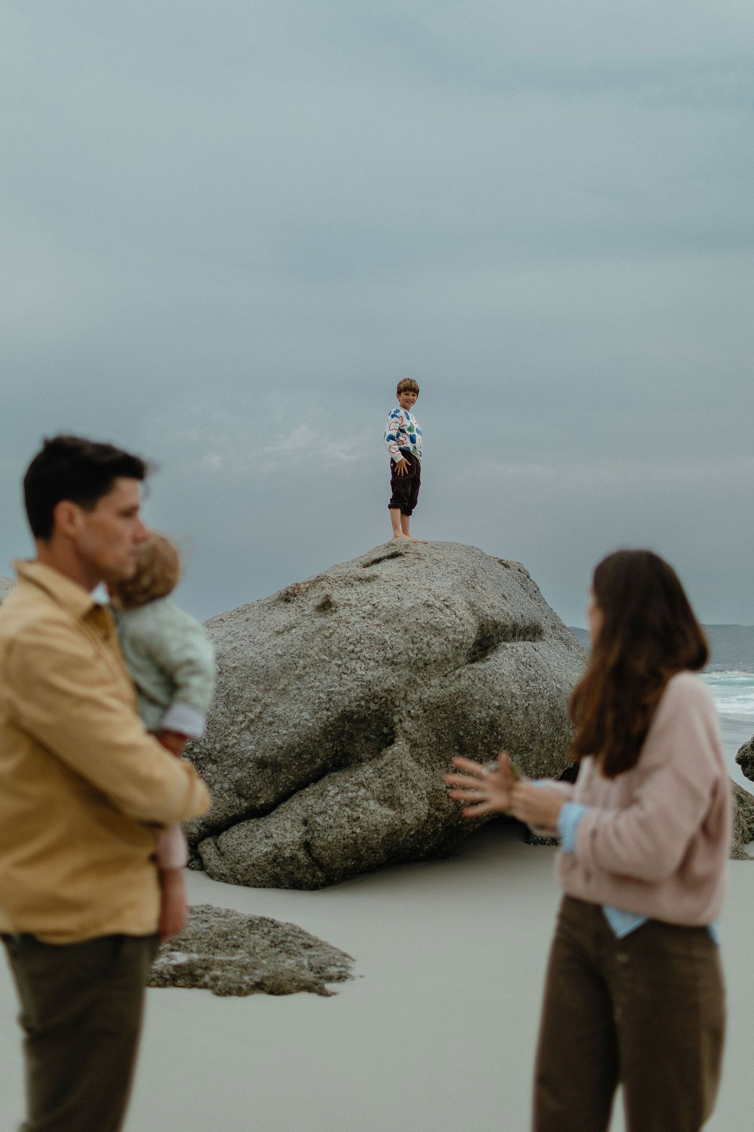 Parents watch from the sandy beach as a young boy stands atop a large boulder under a moody overcast sky.