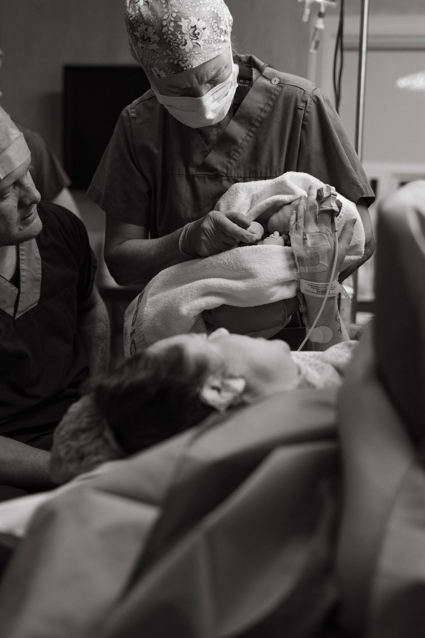 A smiling father and a masked nurse look down at a newborn as the mother lies on the operating table during a cesarean delivery.