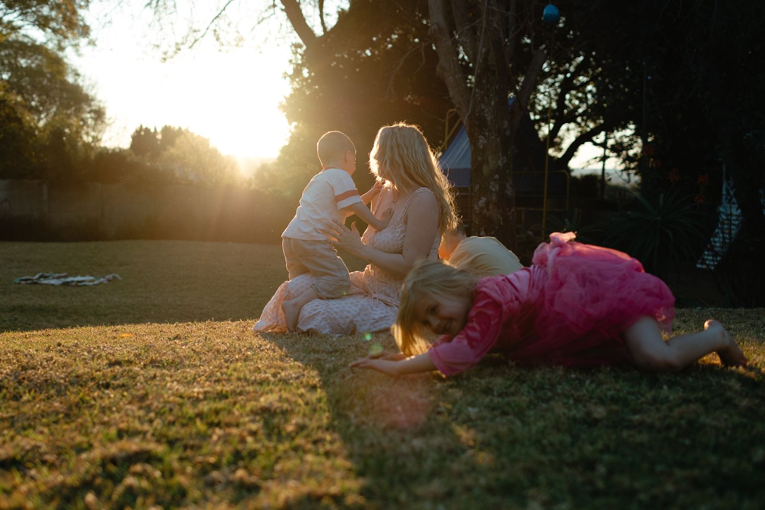 A mother and her children play on a grassy lawn at sunset, with warm sunlight streaming through the trees behind them.