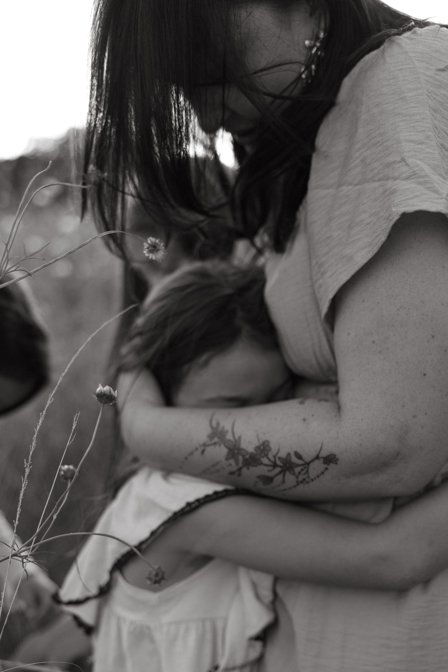 Black and white photo of a mother with a floral arm tattoo tenderly hugging her young daughter in a grassy wildflower field.