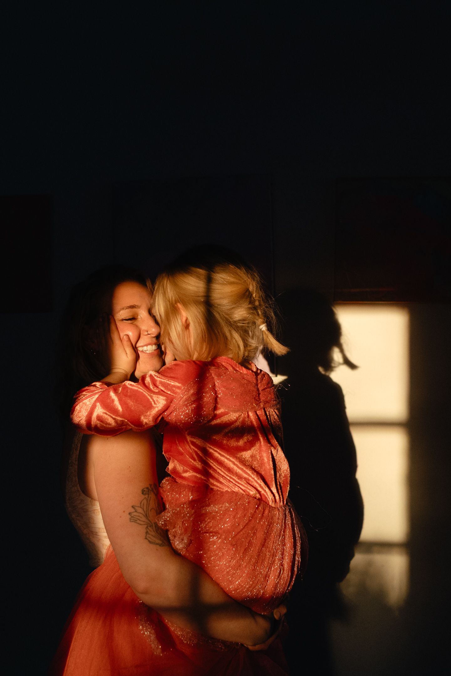 Smiling woman in an orange dress hugs a little girl in a shimmering red outfit in warm, golden light against a dark background.