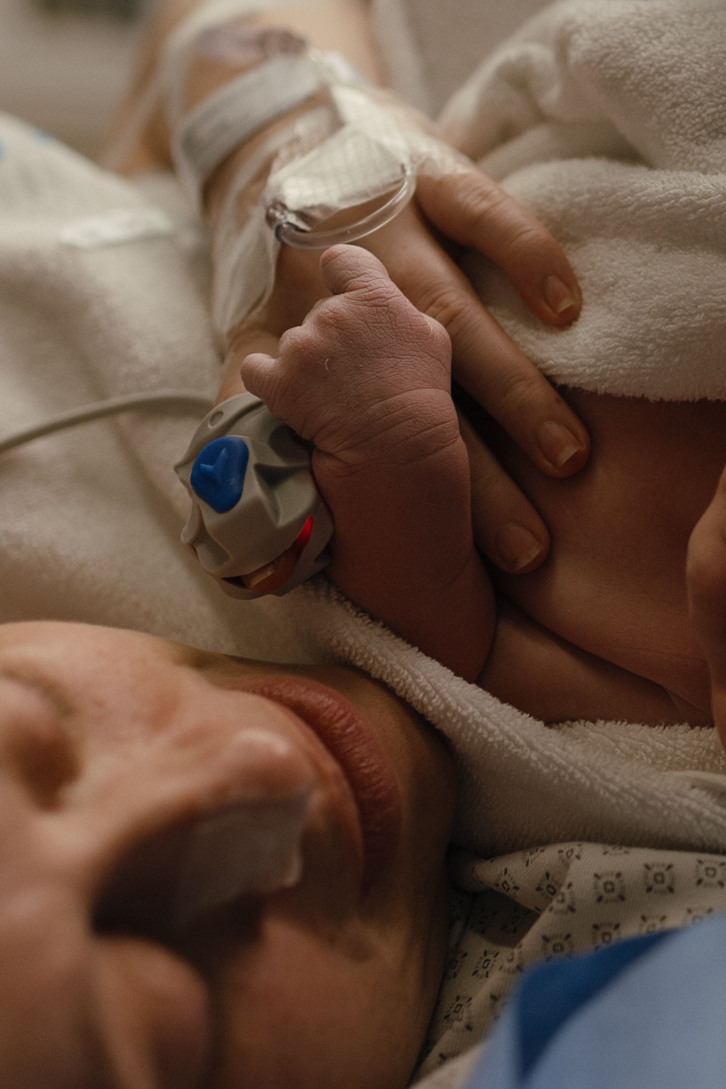 Newborn baby's tiny hand with a hospital ID band rests on a mother's hand as she lies in a hospital bed wrapped in soft white blankets.