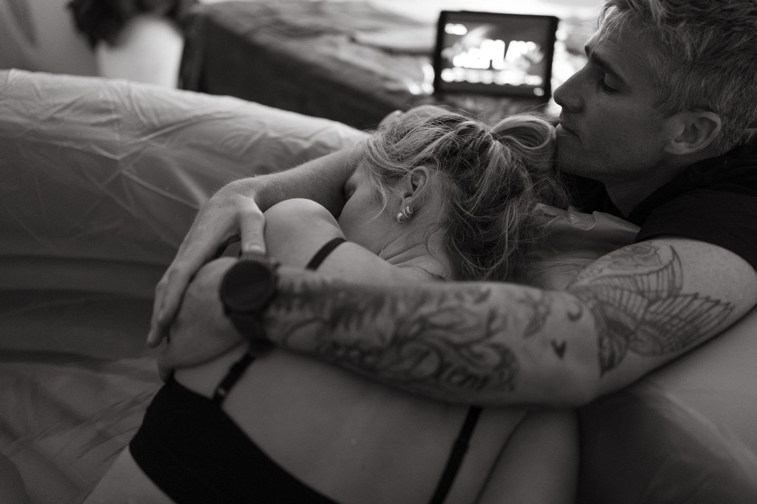 Tattooed partner wraps his arms around a laboring woman, holding her close for support during an intimate birth at home.
