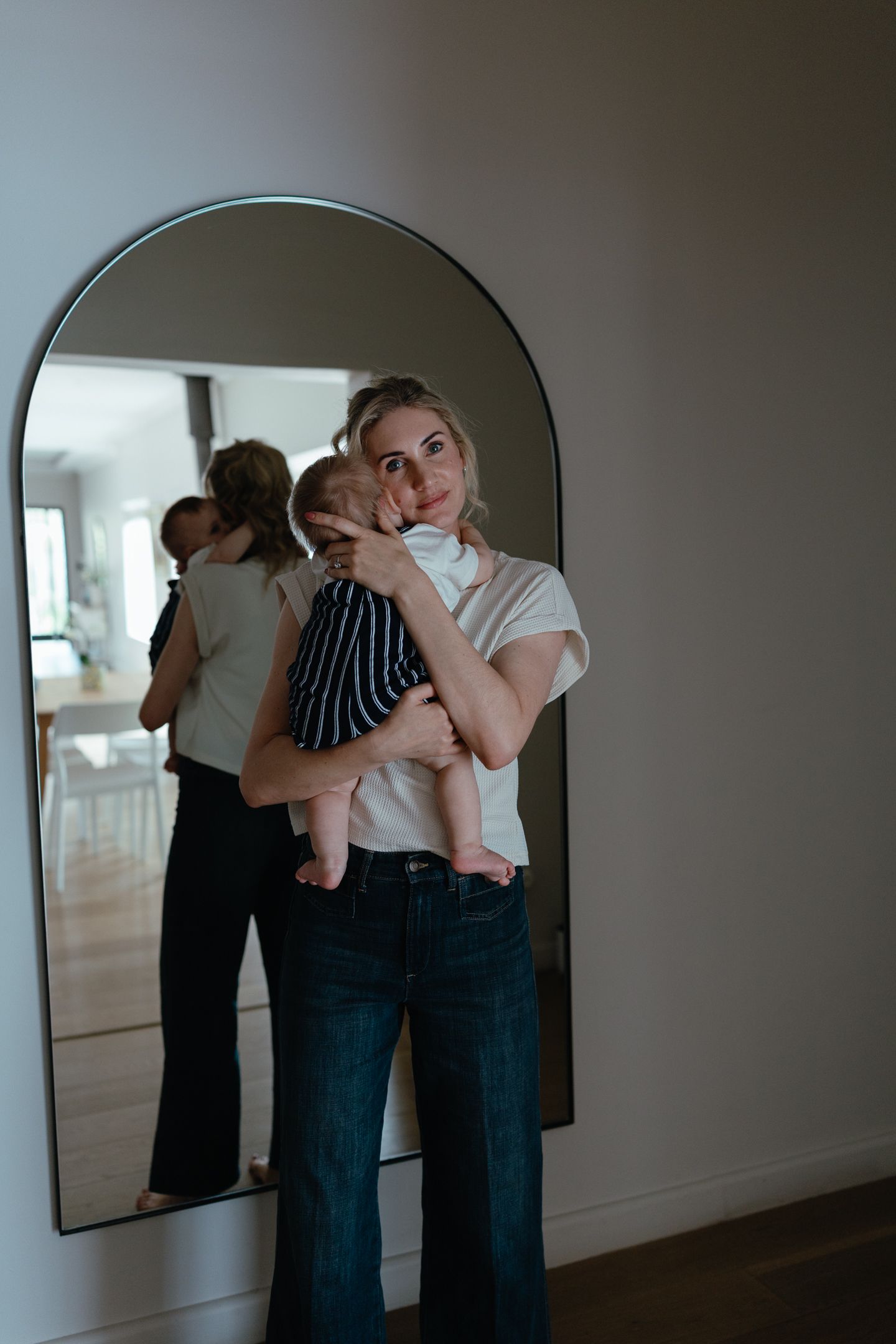 Blonde mother holds her baby on her shoulder in front of a tall arched mirror in a bright home.
