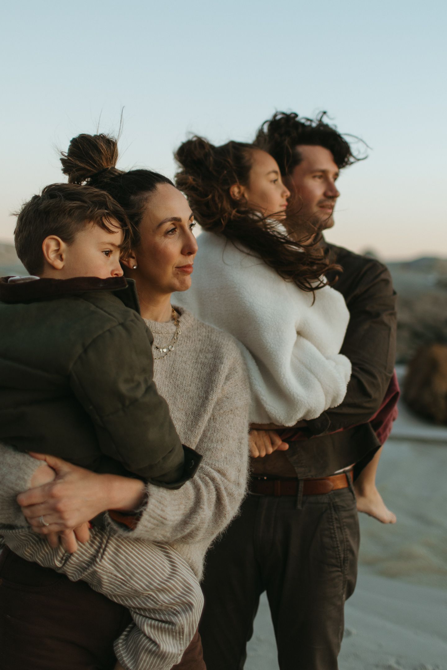A family of four huddles together on the coast at sunset, parents carrying their two children as the wind blows their hair.
