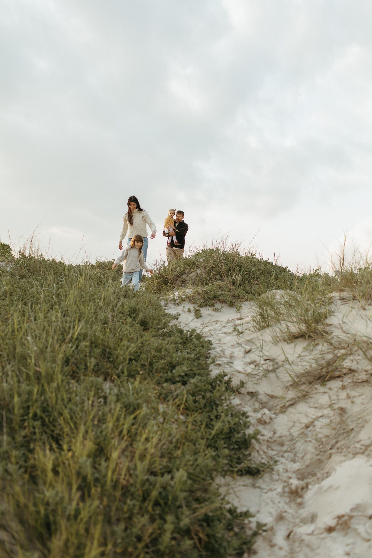 A family of four walks down a grassy sand dune path under an overcast sky near the beach.