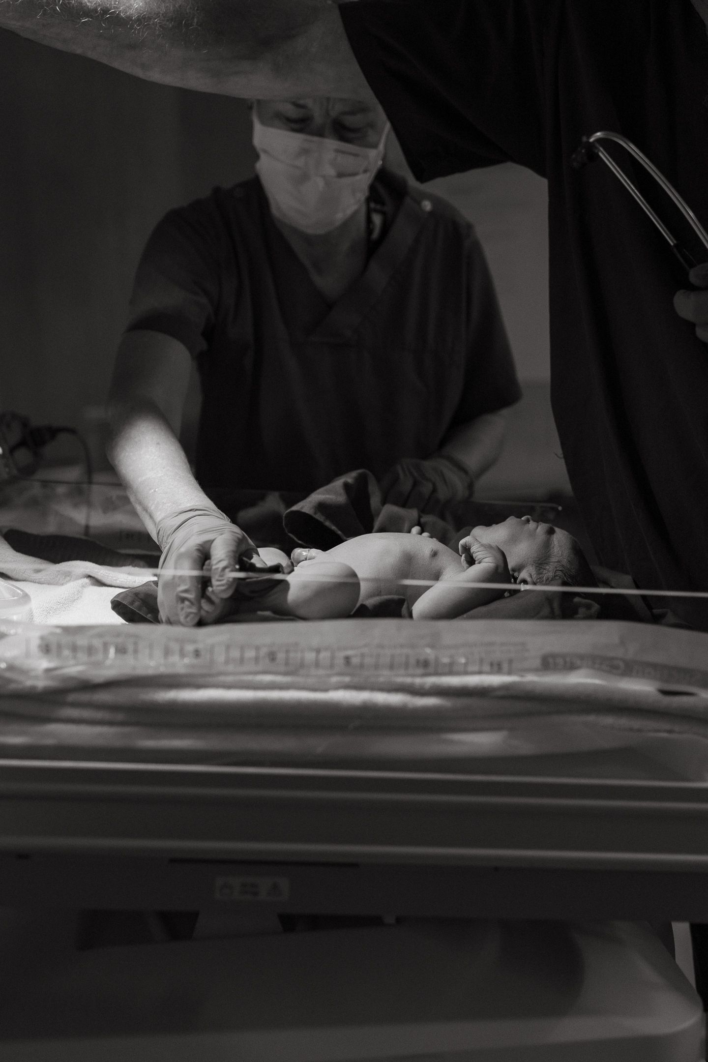 Black and white photo of a masked medical professional in scrubs examining a newborn baby lying in a hospital bassinet.