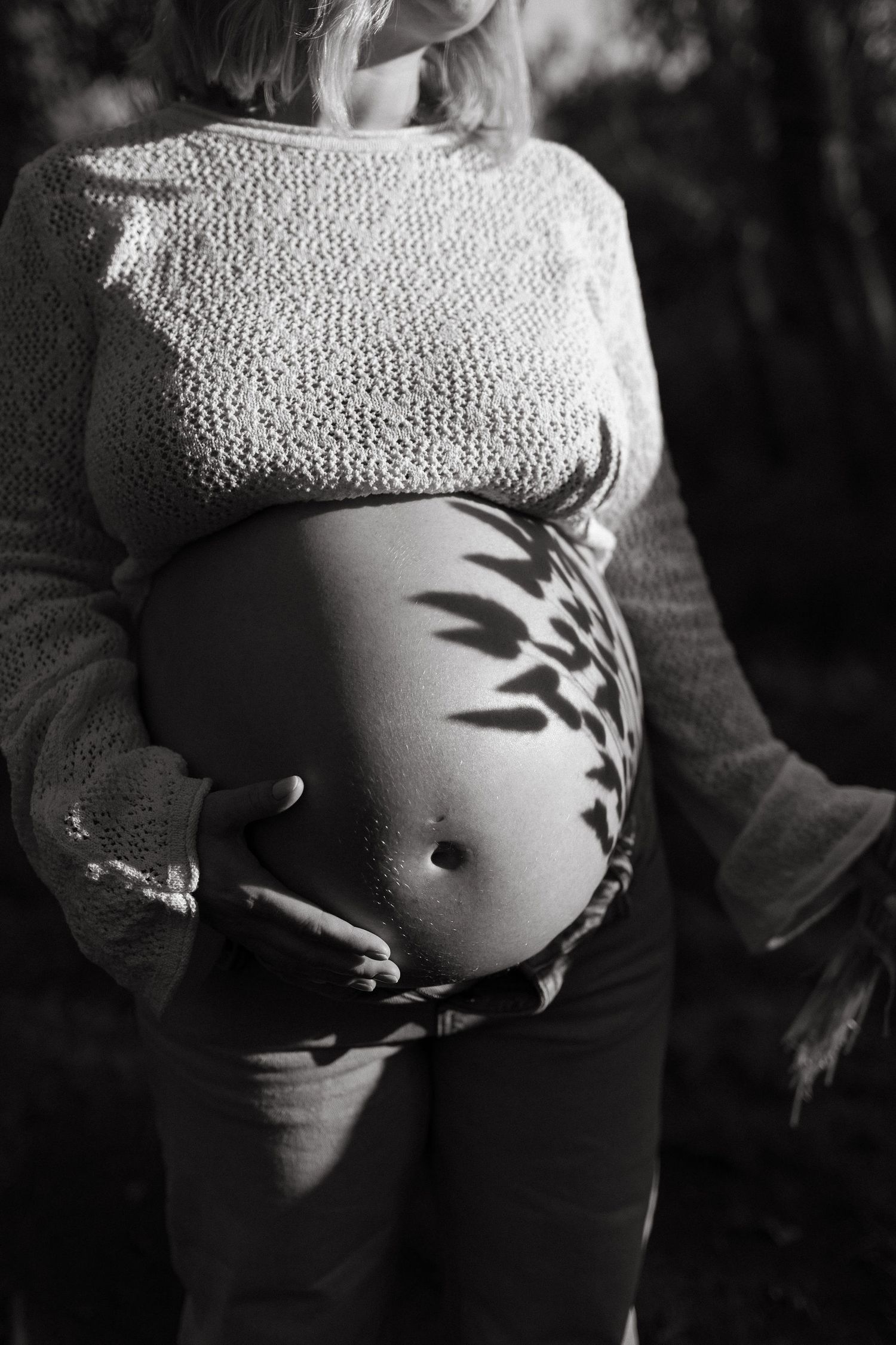 Leaf shadows fall across a pregnant belly as the mother cradles it in a moody black and white maternity photo.