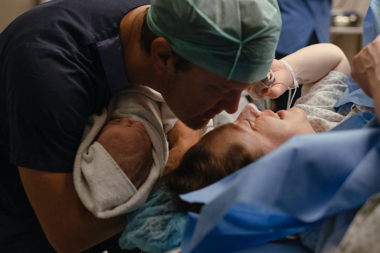 Father in scrubs and surgical cap leans over a mother to kiss her and greet their newborn baby after delivery.