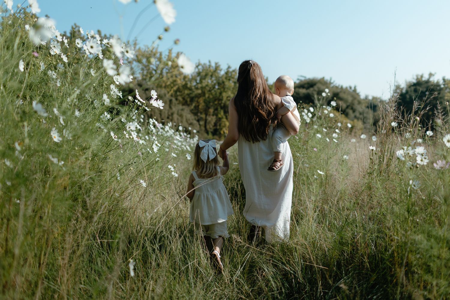 Mother in a white dress walks through a wildflower field holding a baby while a young girl walks beside her.