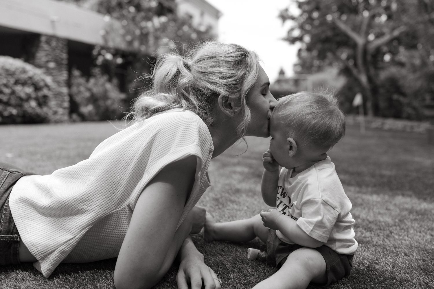 A woman with a ponytail lies on the grass and kisses a toddler sitting beside her in a black and white outdoor photo.