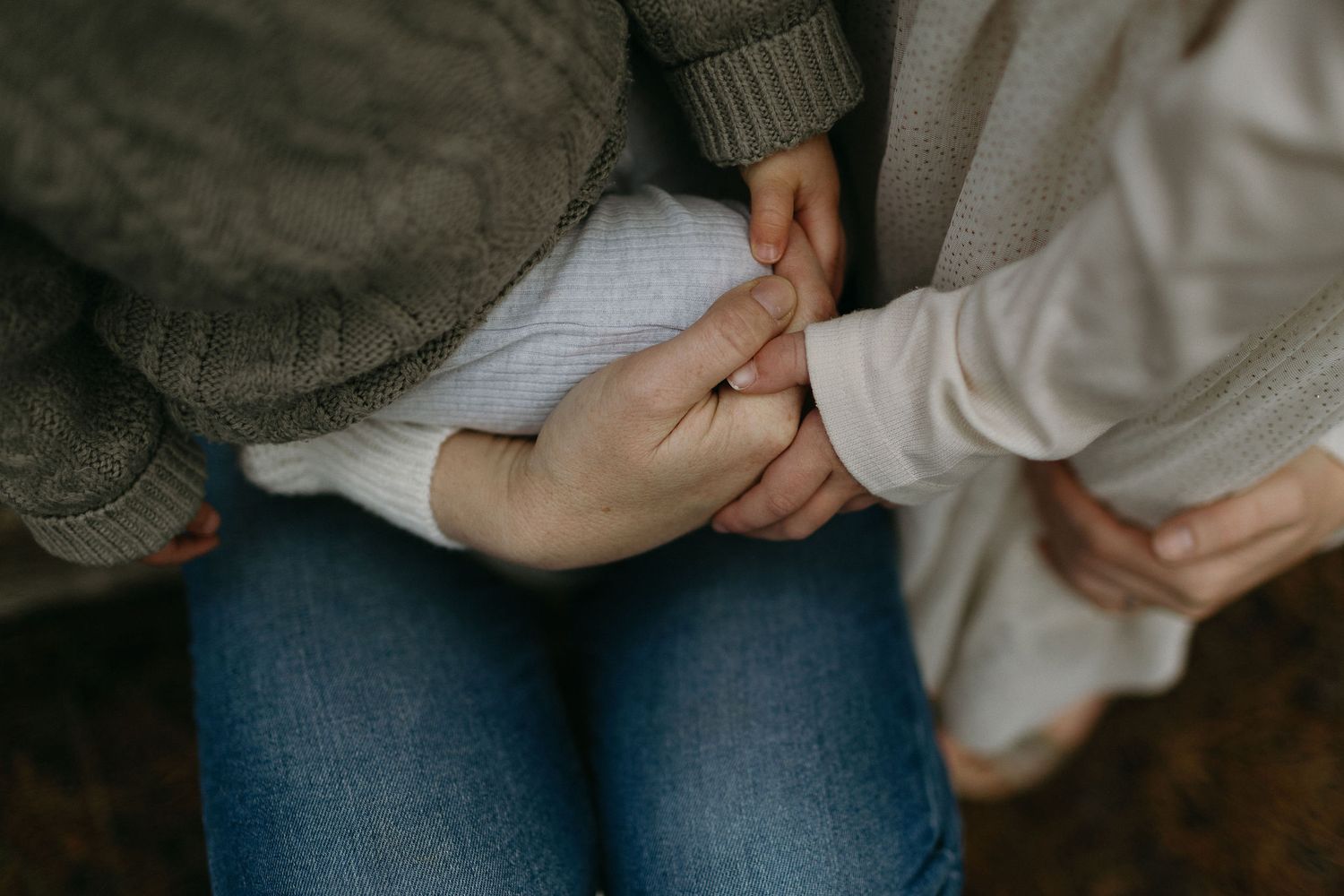 Close-up of a parent's hands gently holding a baby's hands on a lap, wearing jeans and a cozy knit sweater.