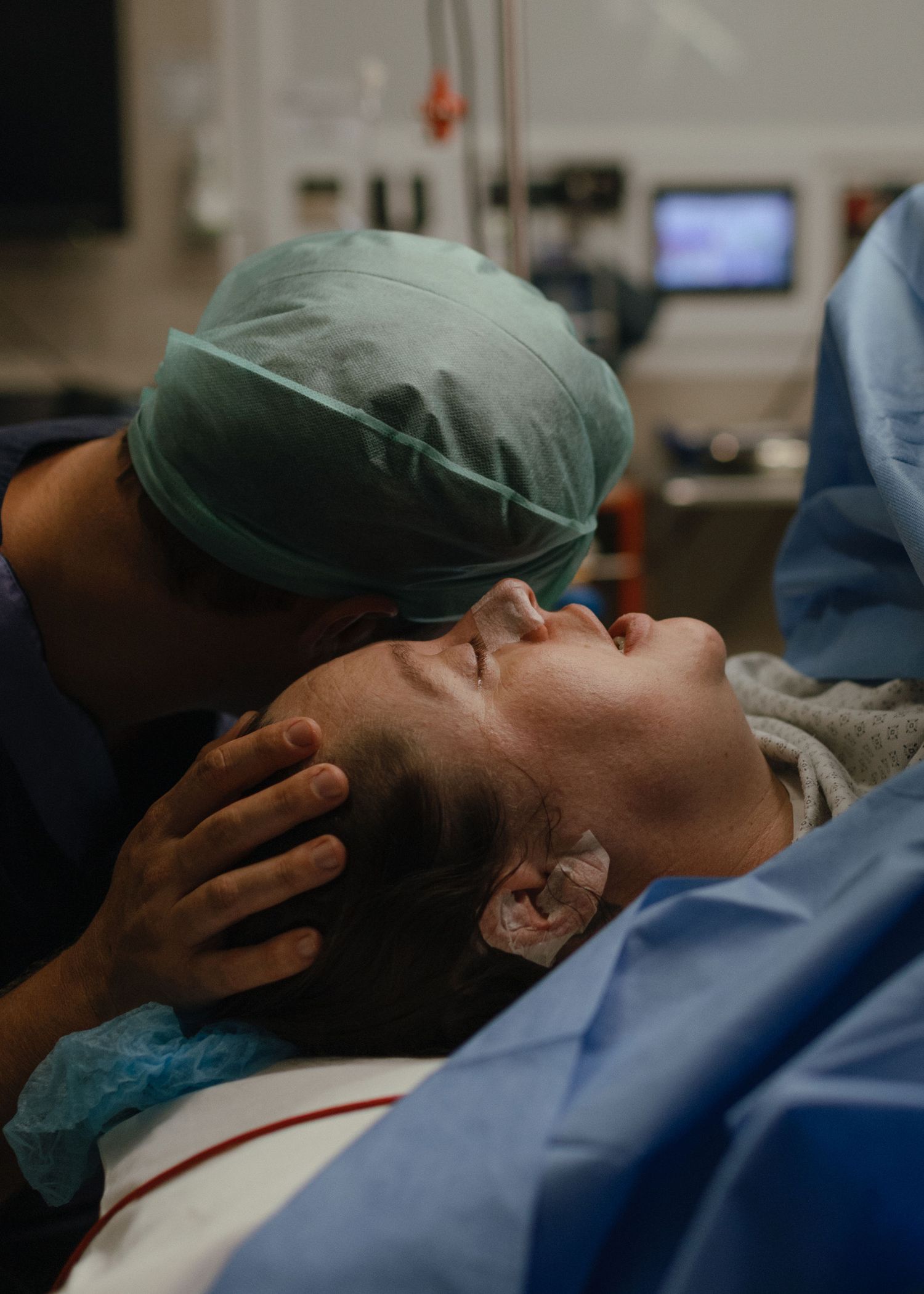 A father in scrubs and a surgical cap tenderly kisses the forehead of the mother lying on the delivery table in an operating room.