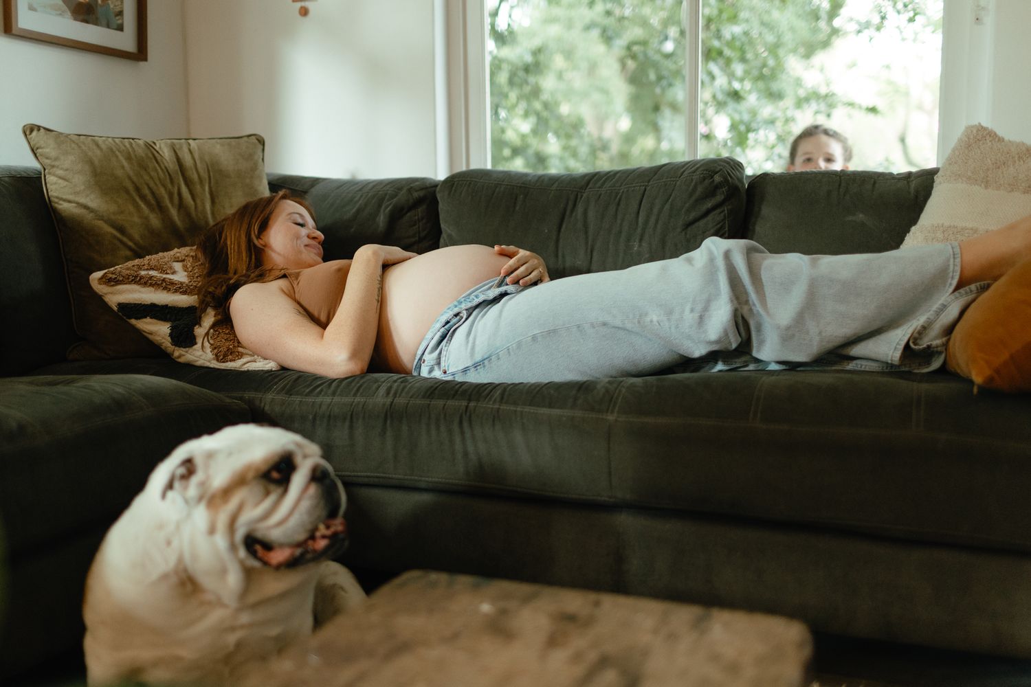 A pregnant woman naps on a green sofa while a sleepy bulldog rests in the foreground of the living room.