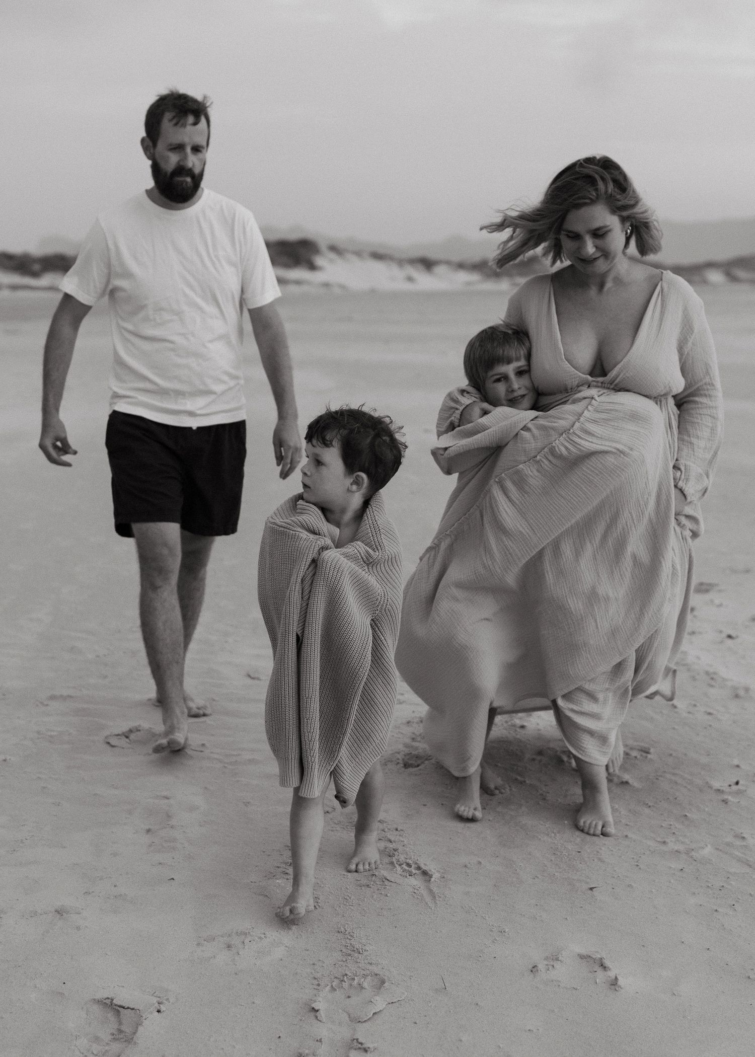 A black and white beach photo of a pregnant mother walking with her husband and two young sons wrapped in blankets.