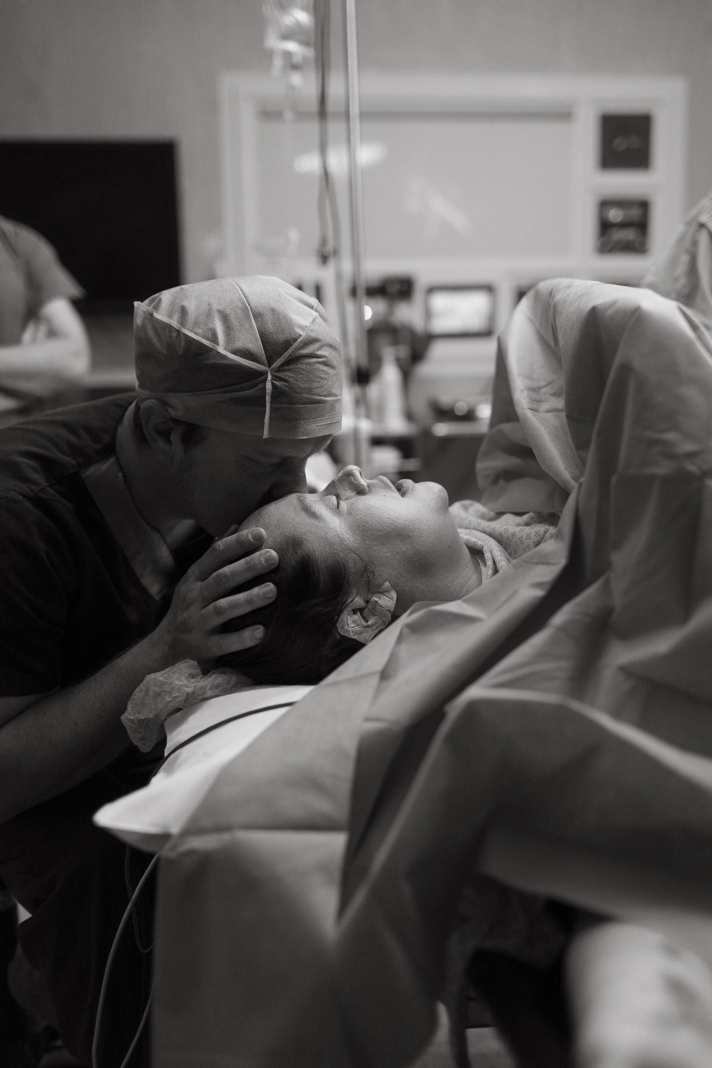 A masked partner in a surgical cap kisses the forehead of a woman lying on an operating table during a cesarean birth, in black and white.